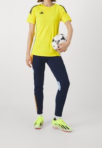 Žluté sportovní tričko s černými akcenty, kombinované s námořnickými kalhotami se žlutými a modrými detaily. Jasně zelené fotbalové kopačky a míč.