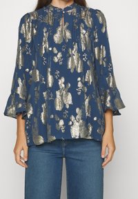 Top azul marino con patrón floral metálico dorado, escote en forma de ojo de cerradura y mangas con volantes. Combinado con jeans de mezclilla azules.