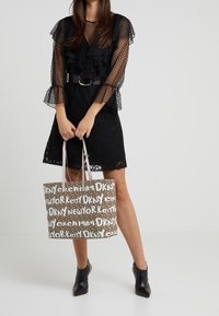 Femme en robe noire en dentelle tenant un sac fourre-tout marron avec le texte blanc "DKNY New York City", portant des bottines à talons noires.