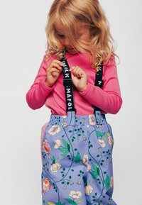 Paarse waterdichte broek met bloemenpatronen en vogels, voorzien van verstelbare zwarte banden met het label "Mafolk." De top is een roze shirt met lange mouwen.