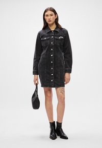 Vestido em denim preto com mangas longas, fecho de botões, dois bolsos no peito e costuras laterais. Modelo segura uma pequena bolsa preta.