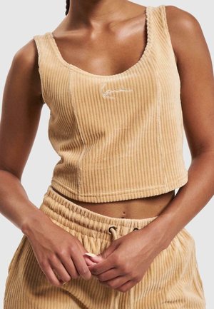 Dámský béžový žebrovaný bezrukávový crop top a odpovídající kalhoty se šňůrkou v pase, ruce sepnuté v pase na světlém pozadí.