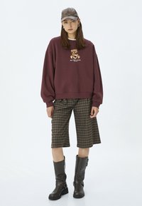 Sudadera color marrón con un gráfico de oso, pantalones cortos bermuda de cuadros y botas altas negras con hebillas. El modelo lleva una gorra marrón.