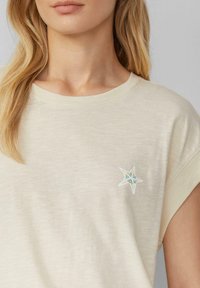 Beige kortemouwen overhemd van zachte stof, met een witte geborduurde ster met blauwe accenten op de linkerborst.