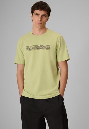 Mężczyzna z krótkimi ciemnymi włosami stoi z rękami w kieszeniach, ubrany w jasnozielony t-shirt z napisem "Catch the Direction" oraz czarne spodnie na szarym tle.