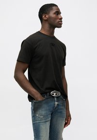 Jovem vestido com uma t-shirt preta com um logotipo, jeans azuis rasgados e um cinto preto com uma fivela prateada, parado com uma mão no bolso.