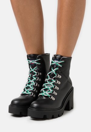 Black leather ankle boots with a chunky heel, featuring green and white laces, silver eyelets, and a rugged sole.