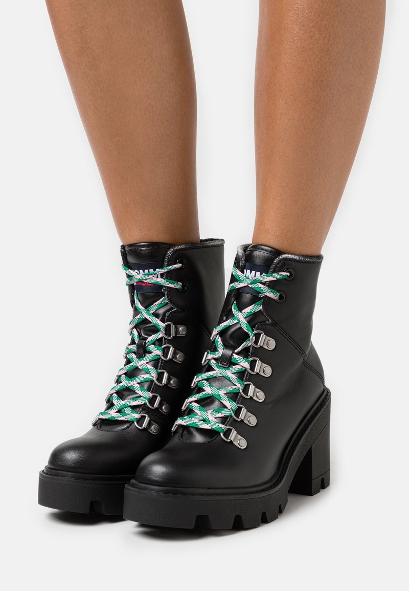 Bottes de cheville en cuir noir avec un talon épais, dotées de lacets verts et blancs, d'œillets argentés et d'une semelle robuste.