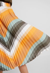 Falda plisada con rayas horizontales en naranja, verde azulado, marrón y blanco, con una tela texturizada que crea profundidad y movimiento.