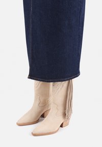 Botas de vaquero de ante beige con punta afilada y diseño en relieve, con flecos en la parte trasera y un tacón de madera apilada, combinadas con jeans oscuros.