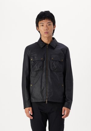 Chaqueta negra con cremallera frontal, dos bolsillos en el pecho y detalles de hardware en bronce. Presenta un collar estructurado y paneles de tela texturizada.