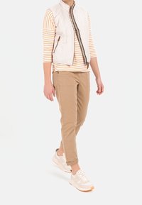 Quiltet hvid ærmeløs vest over en cremet og orange stribet langærmet skjorte, parret med lysebrune bukser og beige sneakers.