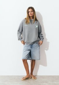 Sweatshirt gris avec un graphique d'étoile, associé à un short en denim clair et des tongs. La tenue présente un design décontracté et relaxé.