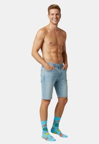 Lichtblauwe denimshorts met een fitted ontwerp, gecombineerd met felleblauwe sokken met een fruitpatroon. Het model heeft kort haar en een neutrale uitdrukking.