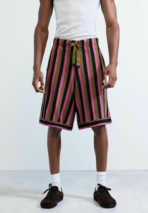 Mężczyzna w wielokolorowych szortach w paski z nadrutką, białych skarpetkach za kostkę, czarnych tenisówkach i białej koszulce bez rękawów, stojący na szarym podłodze.