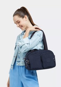 Borsa in tessuto blu navy con due tasche frontali, cerniere argentate e una tracolla staccabile, portata a spalla. Presenta un dettaglio con logo circolare.