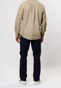 Beige langärmliges Hemd mit Knopfmanschetten, kombiniert mit dunkelblauen Jeans und weißen Sneakers mit roten und blauen Streifen. Glatte Stoffstruktur.