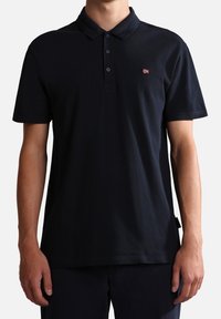 Homme portant un polo noir uni à manches courtes avec trois boutons et un petit logo de drapeau brodé sur la poitrine gauche.