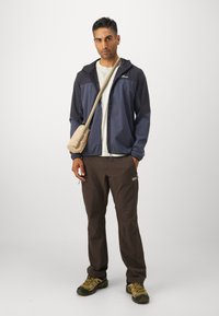Marineblå og grå glidelåsjakke, beige t-skjorte, brune bukser, bærer en liten beige crossover-veske. Har på seg brune og gule tursko.