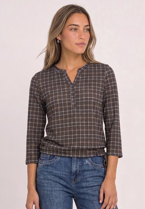 Kvinde med langt blond hår iført brun ternet top med ternede trekvartærmer og blå jeans, kigger til siden mod en ensfarvet baggrund.