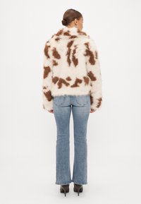 Casaco de pelo sintético em creme com manchas castanhas, apresentando mangas longas e um decote arredondado. Combinado com jeans claros de perna larga e saltos pretos.