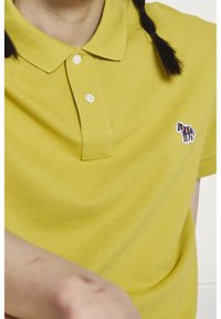 Personne portant un polo jaune avec des boutons blancs et un petit zèbre multicolore brodé sur le côté gauche de la poitrine, cheveux en tresses.