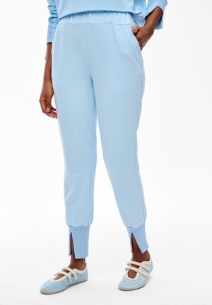 Osoba w jasnoniebieskich spodniach dresowych z rozcięciami przy kostkach i pasujących niebieskich balerinach z beżowymi paskami, stojąca na białym tle.
