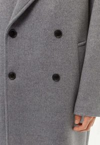 Grå ullkappa med dubbelknäppt design, sex svarta knappar och en texturerad yta med sidoficka och breda slag.