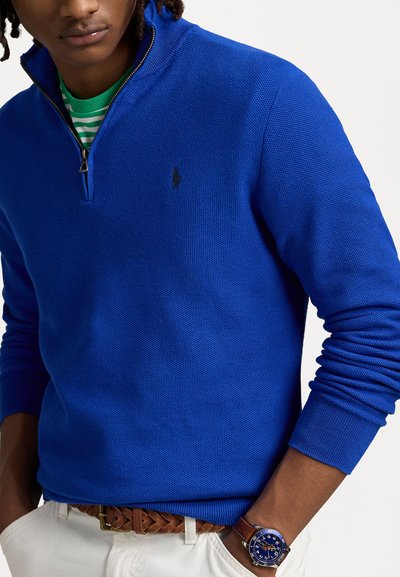 Kraljevo modra zip-up jopica z teksturirano pletenino, z black logotipom na prsih. Obuta nad zeleni in beli črtastim majico.