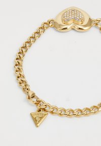 Bracelet en chaîne dorée avec un pendentif en forme de cœur orné de strass et un charm triangulaire marqué "Guess". Texture lisse, finition brillante.
