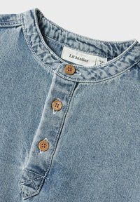 Camicia in denim blu chiaro con scollatura rotonda, dotata di tre bottoni in legno e di una piccola etichetta sul colletto. Tessuto testurizzato, design casual.