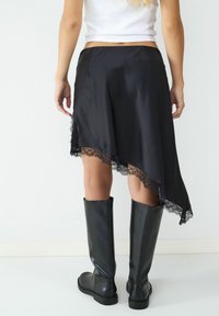 Schwarzer asymmetrischer Rock aus Satin mit einer Spitzenkante. Kombiniert mit knielangen schwarzen Lederstiefeln. Glatte Textur und fließendes Design.