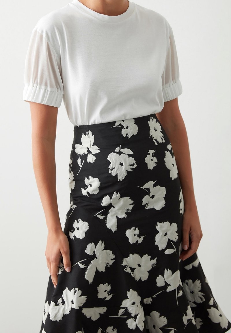 Femme portant un chemisier blanc à manches courtes froncées et une jupe taille haute noire ornée de grands motifs floraux blancs.