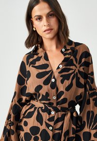 Camicia marrone con bottoni, caratterizzata da un ampio motivo floreale nero, con colletto, maniche lunghe e un dettaglio di vita legato. Tessuto morbido.