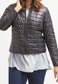 Kviltet sort pufferjakke med frontlynlås, let materiale og cropped design, båret over en lys grå top og blå jeans.