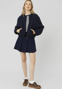 Veste de pilote navy avec une texture pelucheuse, top rayé en dessous, associée à une jupe évasée navy et des chaussures lacées marron avec des chaussettes blanches.
