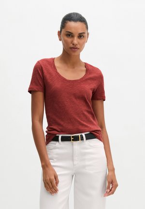 Femme portant un t-shirt rouge rouille à manches courtes rentré dans un pantalon blanc avec une ceinture noire, debout devant un fond blanc uni.