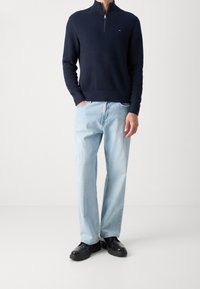Pull en maille bleu marine à fermeture zippée au col, pantalon en denim bleu, et chaussures noires. Le pull présente des poignets côtelés et un petit logo sur la poitrine.