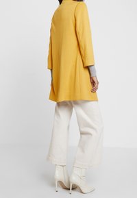 Sininen vaaleankeltainen pitkähihaisten takki, jossa on rento malli, suora leikkaus ja sivuhalkiot, yhdistettynä valkoisiin leveälahkeisiin housuihin ja vaaleanbeigeihin nilkkureihin.