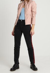 Giacca in ecopelle rosa chiaro con un design aderente, canotta a righe blu e bianche, jeans neri con strisce rosse ai lati e scarpe nere con lacci.