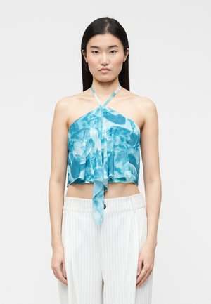 Noor daamede pikkade tumedate juustega naine, kandes sinise lipsuvärviga kaelaümbrise topi ja valge triibulise püksteid, seistes tavalise tausta ees.