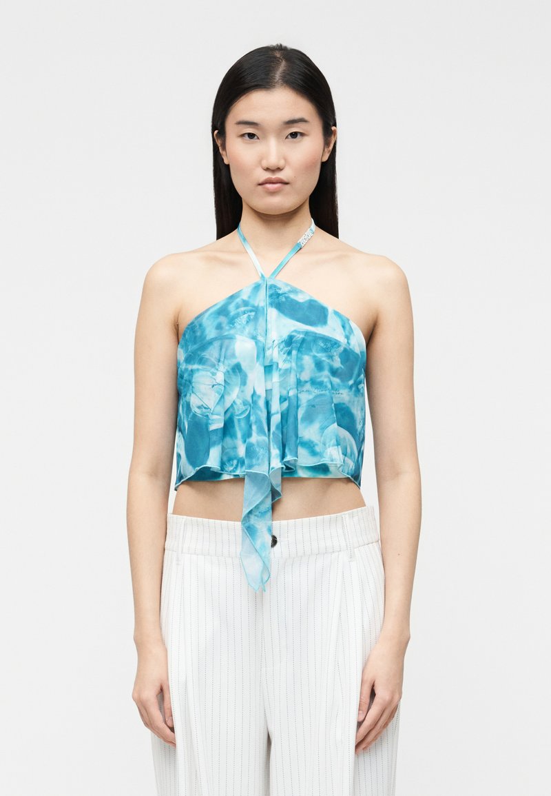 Νεαρή γυναίκα με μακριά σκούρα μαλλιά, ντυμένη με μπλε τοπ με μοτίβο tie-dye και λευκό παντελόνι με ρίγες, στέκεται μπροστά σε απλό φόντο.