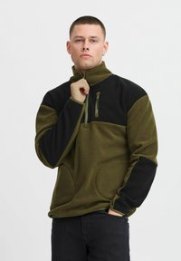 Fleece genser i olivengrønn og svart, med glidelås i kragen, brystlomme og sidelommer med en glatt tekstur.