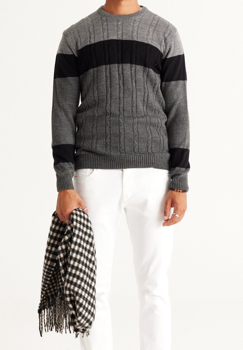 Homme portant un pull en maille torsadée gris avec des rayures horizontales noires, pantalon blanc, tenant une écharpe à carreaux noir et blanc dans la main gauche.