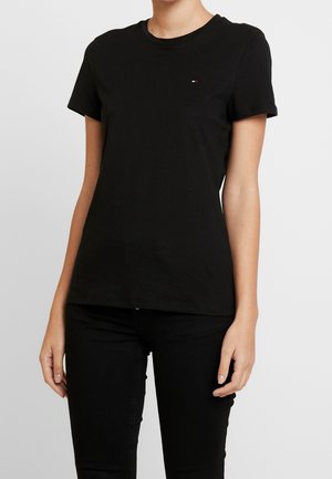 Femme portant un t-shirt noir uni à manches courtes et un pantalon noir, debout devant un fond uni gris clair.