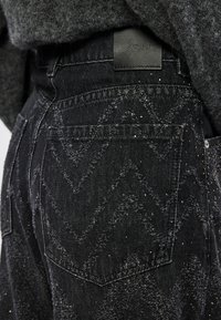 Jeans de denim preto com um padrão zigzag e suaves brilhos, com um patch de marca em couro e bolsos padrão. A textura parece ligeiramente desgastada.