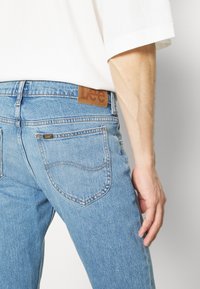 Jeans de mezclilla azul con cintura alta, que presentan una etiqueta de cuero marrón en la parte trasera. La tela muestra una textura desgastada con bolsillos cosidos.