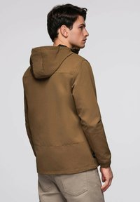 Jeune homme de profil, tourné vers la gauche, portant une veste à capuche marron et un pantalon beige, le tout sur un fond gris clair uni.