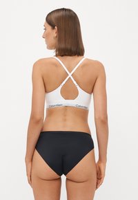Witte sportbeha met een sleutelgatsnede, gekruiste banden en een zwarte logoband. Zwarte bikini onderkant met een gladde textuur en hoge uitsnede.