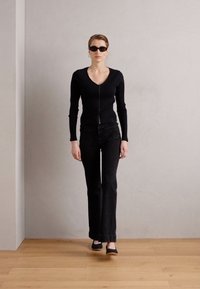 Crna rebrasta bluza dugih rukava s patentnim zatvaračem, uparena s crnim trapericama visokog struka. Model nosi tamne naočale i crne balerinke na drvenom podu.